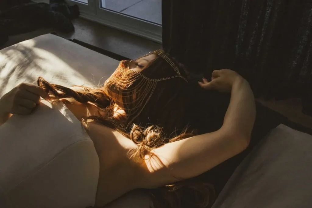 Ava Marie bathed in a ray of sunlight shining through the window, stretched out on her back with her hair fanning out around her. She held a dainty chain headpiece over her eyes, creating an atmosphere of peace and contemplation. Ava Marie Halifax's Elite Independent Companion